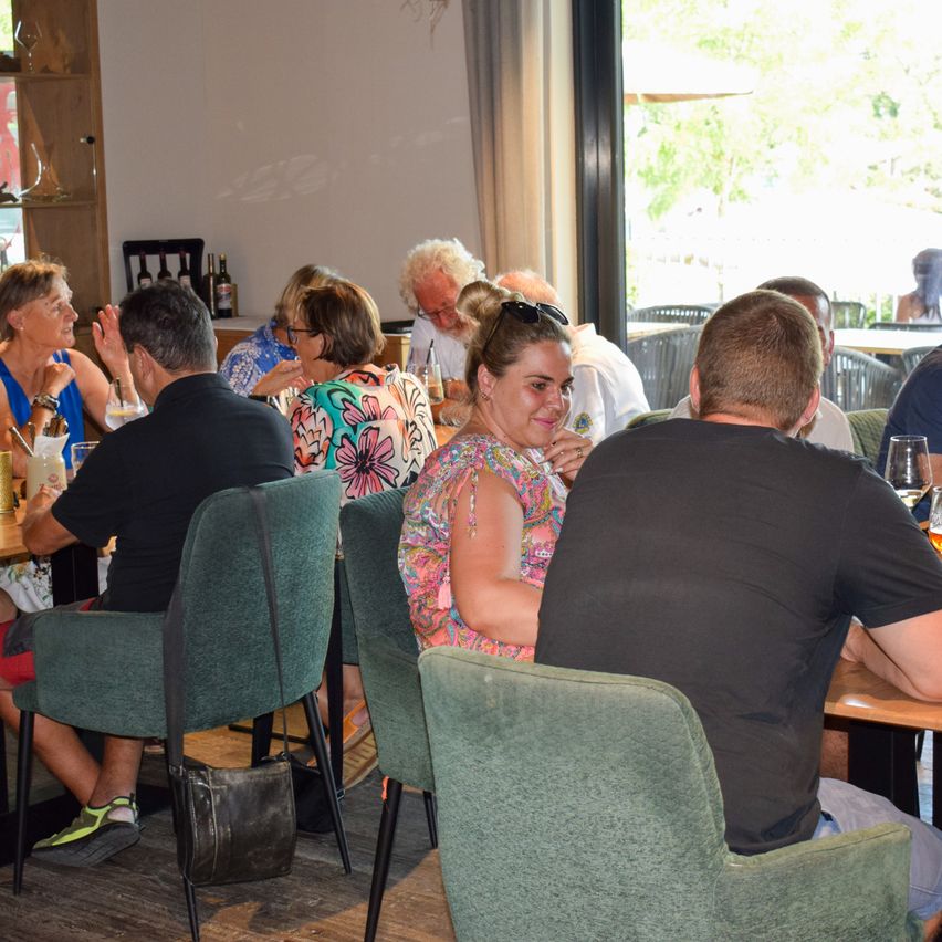 Eine Gruppe von Menschen sitzt an einem Tisch in einem Restaurant. Einige schauen nach vorne, während andere in ein Gespräch vertieft sind. Der Tisch ist mit Gläsern, Tellern und Weinflaschen gedeckt.