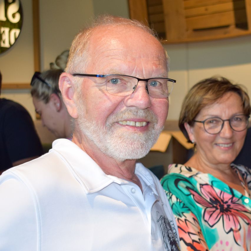 Ein älterer Mann mit Brille und Bart lächelt neben einer Frau mit Brille und einem Blumenshirt. Im Hintergrund sehen wir verschwommen andere Leute.