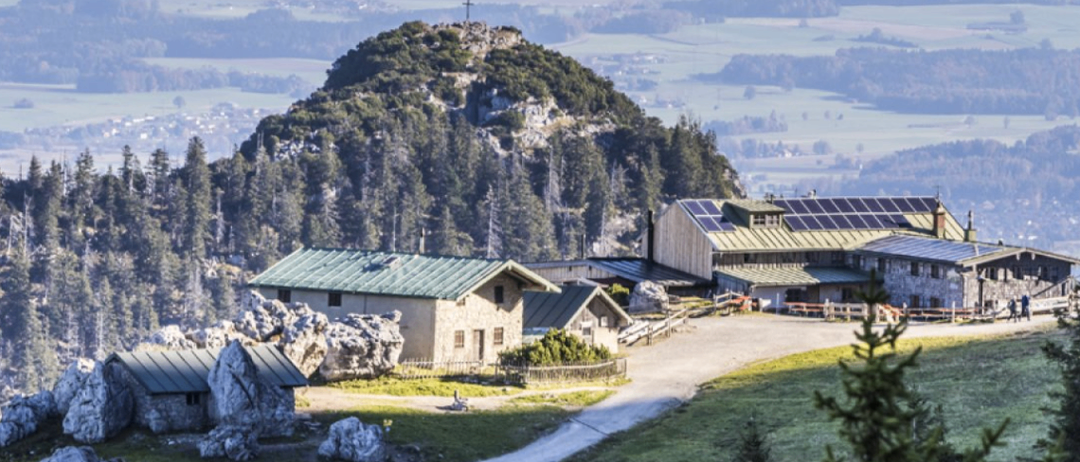 Luftaufnahme eines Berggipfels mit einem Kreuz, umgeben von Bäumen. Darunter ist ein Haus mit Solarpaneelen zu sehen, zusammen mit einem Gebäude. Das Gebiet ist mit großen Felsen gefüllt und hat eine Fernsicht auf eine Stadt und ein Tal.