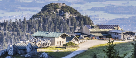 Luftaufnahme eines Berggipfels mit einem Kreuz, umgeben von Bäumen. Darunter ist ein Haus mit Solarpaneelen zu sehen, zusammen mit einem Gebäude. Das Gebiet ist mit großen Felsen gefüllt und hat eine Fernsicht auf eine Stadt und ein Tal.