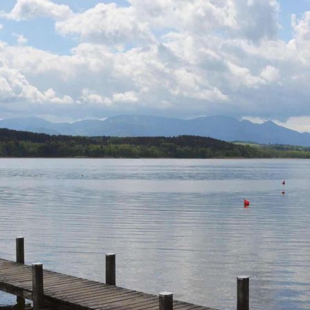Ein ruhiger See mit einem hölzernen Steg erstreckt sich ins Wasser, flankiert von Bergen unter einem bewölkten Himmel.