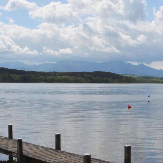 Ein ruhiger See mit einem hölzernen Steg erstreckt sich ins Wasser, flankiert von Bergen unter einem bewölkten Himmel.