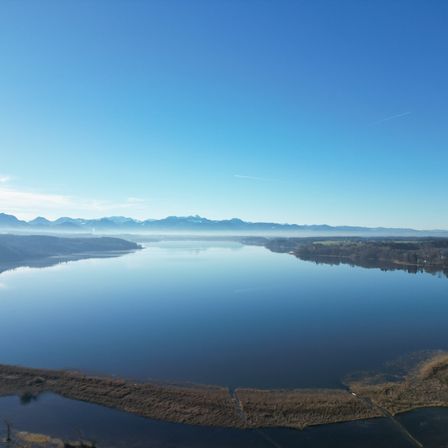Ein ruhiger Luftblick auf einen großen See mit klarem blauem Himmel, entfernten Bergen und einer kleinen Insel im Vordergrund.