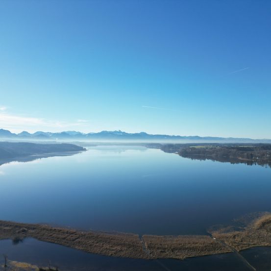 Ein ruhiger Luftblick auf einen großen See mit klarem blauem Himmel, entfernten Bergen und einer kleinen Insel im Vordergrund.