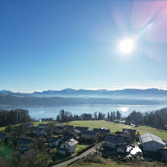 Luftaufnahme eines ruhigen Dorfes mit Häusern, einem See und Bergen im Hintergrund unter einem strahlenden Sonnenschein.