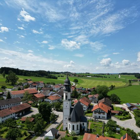 Ein Luftbild eines kleinen Dorfes mit einem markanten Kirchturm, umgeben von üppigen grünen Feldern und einem klaren blauen Himmel.