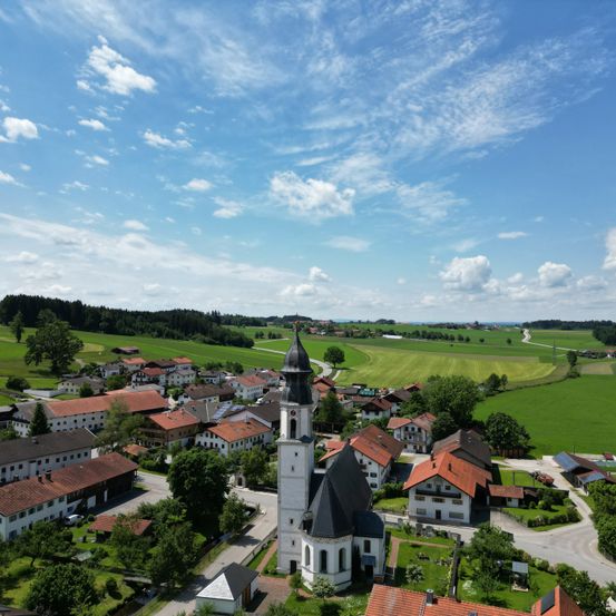 Ein Luftbild eines kleinen Dorfes mit einem markanten Kirchturm, umgeben von üppigen grünen Feldern und einem klaren blauen Himmel.