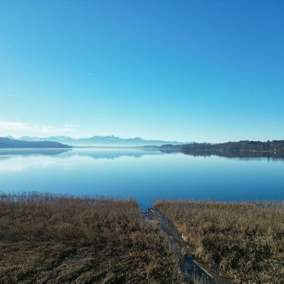 Ein ruhiger See mit ruhigen Wassern, die den blauen Himmel und die entfernten Berge unter einem klaren Himmel widerspiegeln.