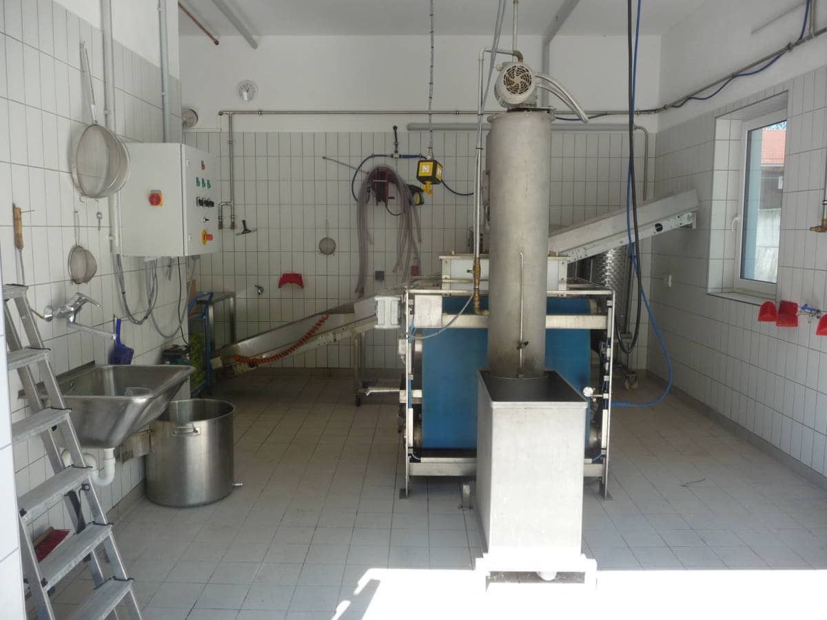 Eine industrielle Küche mit Edelstahlgeräten und gefliesten Böden. Ein großer Metallbehälter und eine hohe zylindrische Maschine stehen in der Mitte.