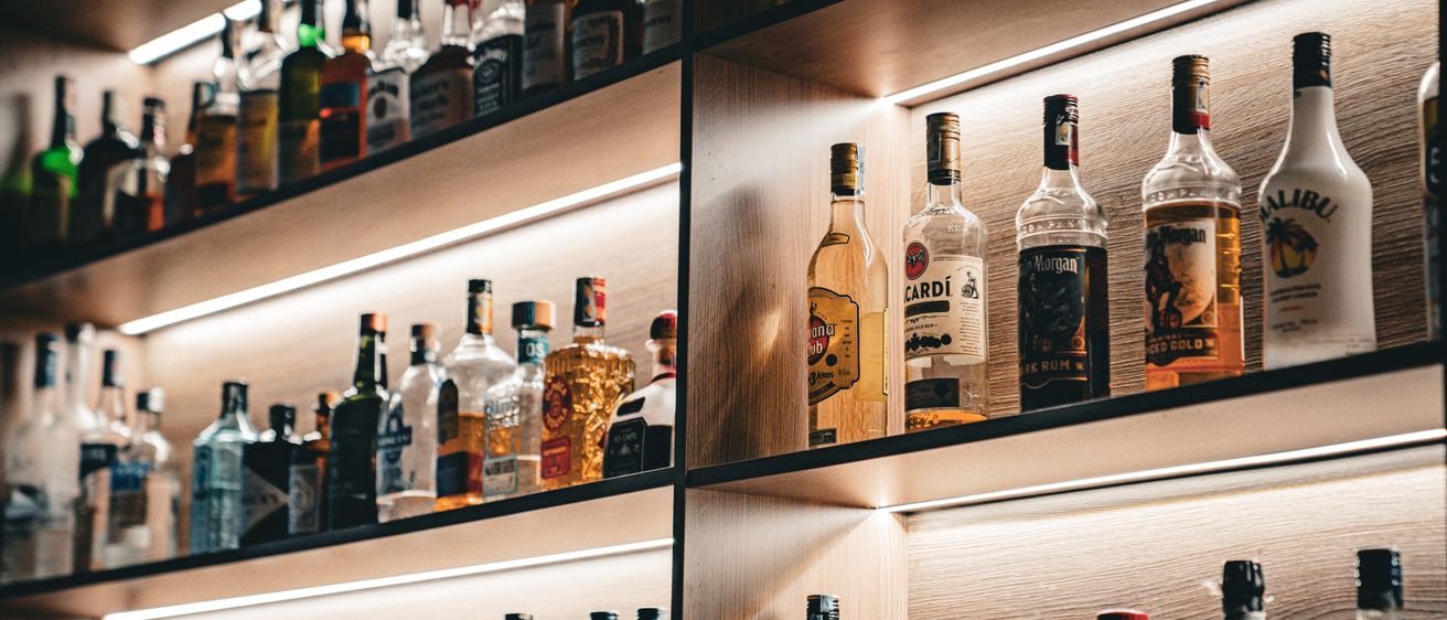 Ein Holzregal mit verschiedenen Likörflaschen, beleuchtet von einem Licht, zeigt die oberen und unteren Regale einer Bar.