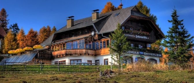 Ein großes Haus mit vielen Fenstern und einem Balkon, es hat Solarpanele und einen Zaun. Bäume umgeben das Haus.