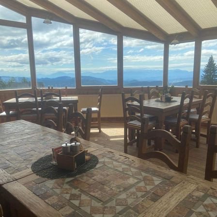 Ein Restaurant mit Holztischen und Stühlen ist zu sehen. Es hat große Fenster mit Blick auf eine Bergkette mit blauem Himmel.