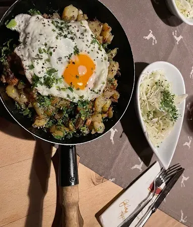 Eine Bratpfanne mit einem Spiegelei auf gebratenen Kartoffeln. Rechts ist eine Schüssel mit Salat. Besteck liegt auf einem Serviettentuch auf dem Tisch.