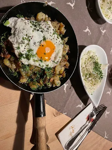 Eine Bratpfanne mit einem Spiegelei auf gebratenen Kartoffeln. Rechts ist eine Schüssel mit Salat. Besteck liegt auf einem Serviettentuch auf dem Tisch.