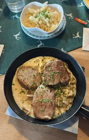 Eine schwarze Bratpfanne auf einem Holztisch enthält ein Gericht mit cremiger Sauce und Fleischscheiben, garniert mit Kräutern. Daneben steht eine weiße Schüssel mit Nudeln und ein Glas Wasser.