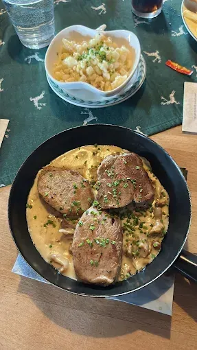 Eine schwarze Bratpfanne auf einem Holztisch enthält ein Gericht mit cremiger Sauce und Fleischscheiben, garniert mit Kräutern. Daneben steht eine weiße Schüssel mit Nudeln und ein Glas Wasser.