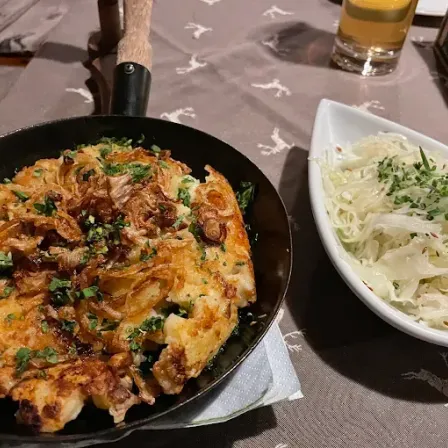 Eine Bratpfanne mit einem goldbraunen Gericht, das mit Kräutern und karamellisierten Zwiebeln garniert ist, steht auf einem Tisch mit einer weißen Schüssel Sauerkraut.