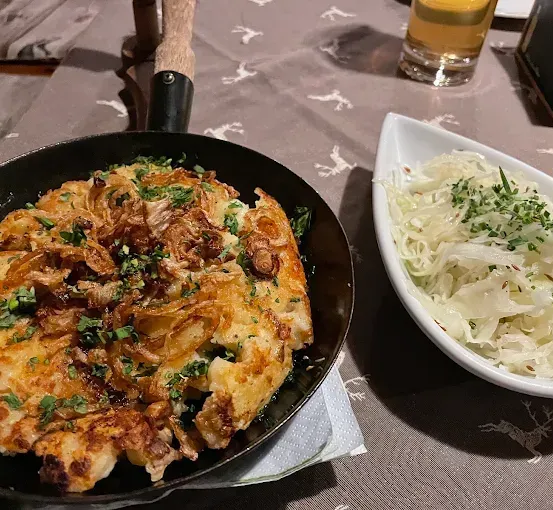 Eine Bratpfanne mit einem goldbraunen Gericht, das mit Kräutern und karamellisierten Zwiebeln garniert ist, steht auf einem Tisch mit einer weißen Schüssel Sauerkraut.