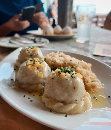 Ein Teller mit Knödeln, Sauerkraut und Sauce steht auf einem Holztisch. Eine Person sitzt am Tisch und schreibt eine SMS.