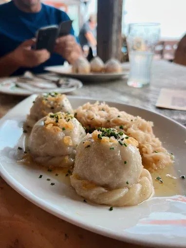 Ein Teller mit Knödeln, Sauerkraut und Sauce steht auf einem Holztisch. Eine Person sitzt am Tisch und schreibt eine SMS.