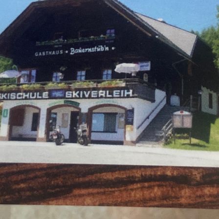 Eine Holzvisitenkarte mit dem Namen 'Gasthaus-Bauernstubn' und den Telefonnummern 3928 und 339. Sie zeigt auch ein Bild eines Skischulgebäudes mit zwei vor dem Gebäude geparkten Motorrädern.