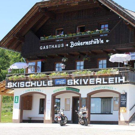 Ein Holzgebäude mit Balkon und der Aufschrift 'Gasthaus Bauernstube' oben. Zwei Motorräder vor dem Gebäude. Eine Treppe führt zum Eingang. Das Schild 'SCHULE SKIVERLEIH' befindet sich am Gebäude.