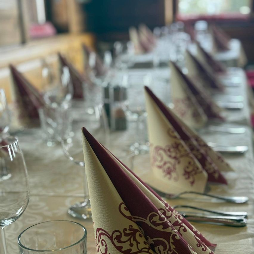 A dining table is set with multiple wine glasses and plates. Each place setting includes a folded napkin with intricate designs. Silverware is neatly arranged beside each setting. The table is covered with a patterned tablecloth. In the background, a blurred view of a window and wall can be seen.