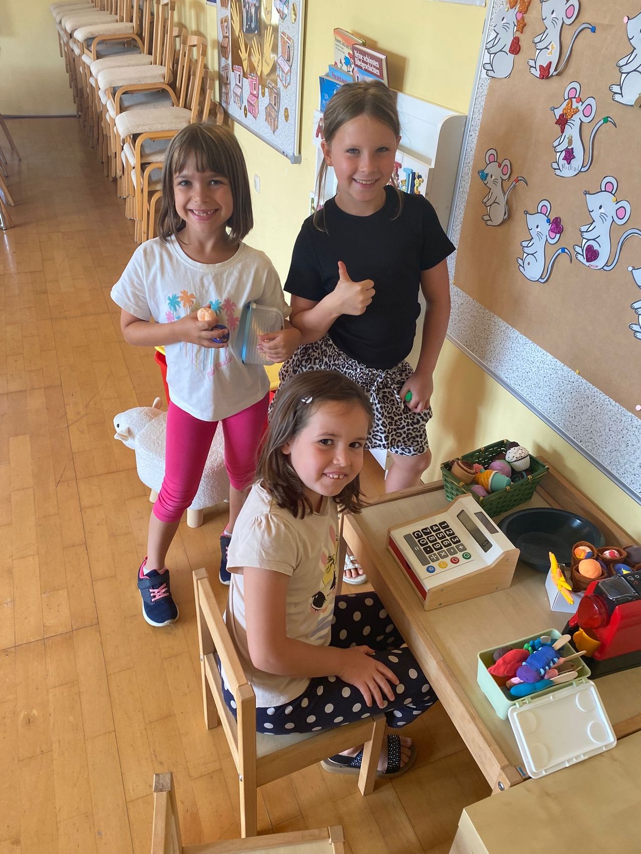 Drei junge Mädchen spielen in einem Klassenzimmer. Eine gibt einen Daumen hoch, eine andere hält ein Spielzeug und die dritte sitzt an einem Schreibtisch mit einer Kasse.