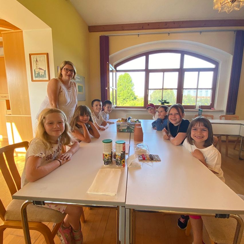 Eine Gruppe von Kindern und einer erwachsenen Frau sitzen an einem Tisch in einem Esszimmer. Auf dem Tisch befinden sich verschiedene Gegenstände. Eine Frau steht hinter ihnen in der Nähe eines Fensters.