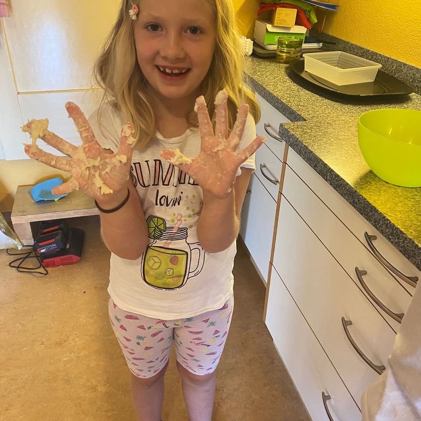 Ein junges Mädchen mit blonden Haaren lächelt in einer Küche, ihre Hände sind mit Mehl bedeckt. Sie trägt ein weißes T-Shirt mit einem Sommer-Design und rosa Shorts. Hinter ihr steht eine Arbeitsplatte mit einer grünen Schüssel und verschiedenen Gegenständen.