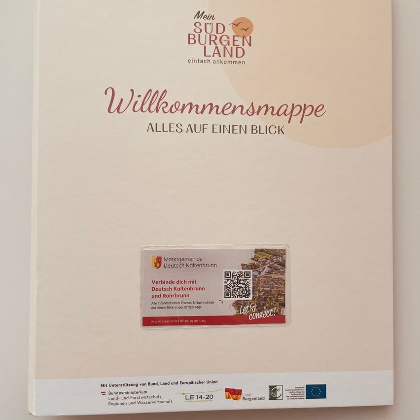 Ein weißes Buch mit dem Titel 'Mein Südburger Land Willkommensmappe Alles auf einen Blick'. Unter dem Titel befindet sich ein quadratischer Aufkleber mit einem QR-Code und Text in Rot und Weiß. Unter dem Aufkleber befindet sich ein Bild einer Stadt. Der untere Teil des Buches hat Logos und Text.