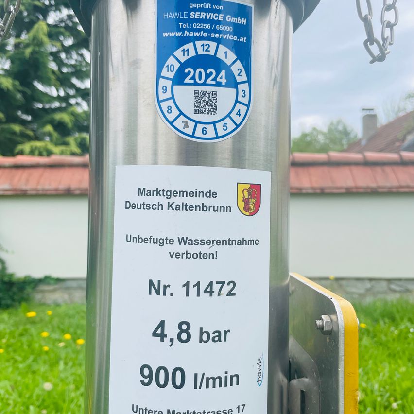 Ein Metallpfosten mit einem blauen Aufkleber zeigt Informationen zu Wassernutzungsbeschränkungen. Der Aufkleber hat ein Logo, einen QR-Code und die Nummer 11472. Er zeigt einen Druck von 4,8 bar und eine Durchflussrate von 900 Litern pro Minute an. Der Standort ist Untere Marktstrasse 17.