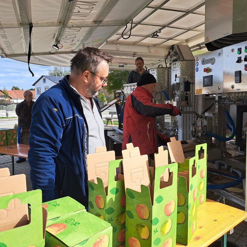 Ein Mann in einer blauen Jacke beobachtet einen Mann in einer roten Jacke, der grüne Kisten mit Äpfeln füllt. Die Kisten sind auf einem Tisch unter einem Vordach angeordnet. Eine Maschine mit einem Schlauch ist in der Nähe.