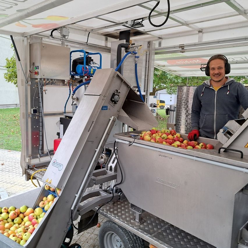 Ein Mann in einer grauen Jacke steht neben einer Maschine, die Äpfel sortiert, mit einem Haufen Äpfel in einem Edelstahlbehälter. Die Maschine hat ein Förderband und ist mit einem Schlauch verbunden. Hinter der Maschine stehen ein Baum und ein Gebäude.