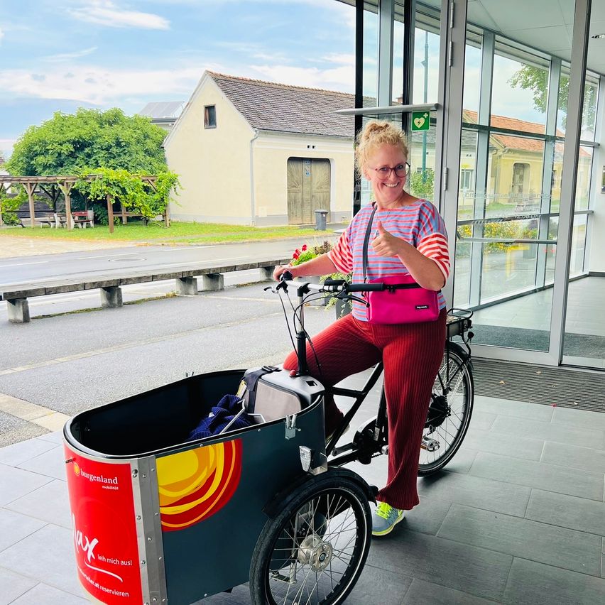 Eine Frau fährt ein Dreirad mit einem Korb vorne, trägt eine Brille und ein buntes gestreiftes Shirt. Das Dreirad hat ein rotes und gelbes Design an der Seite.