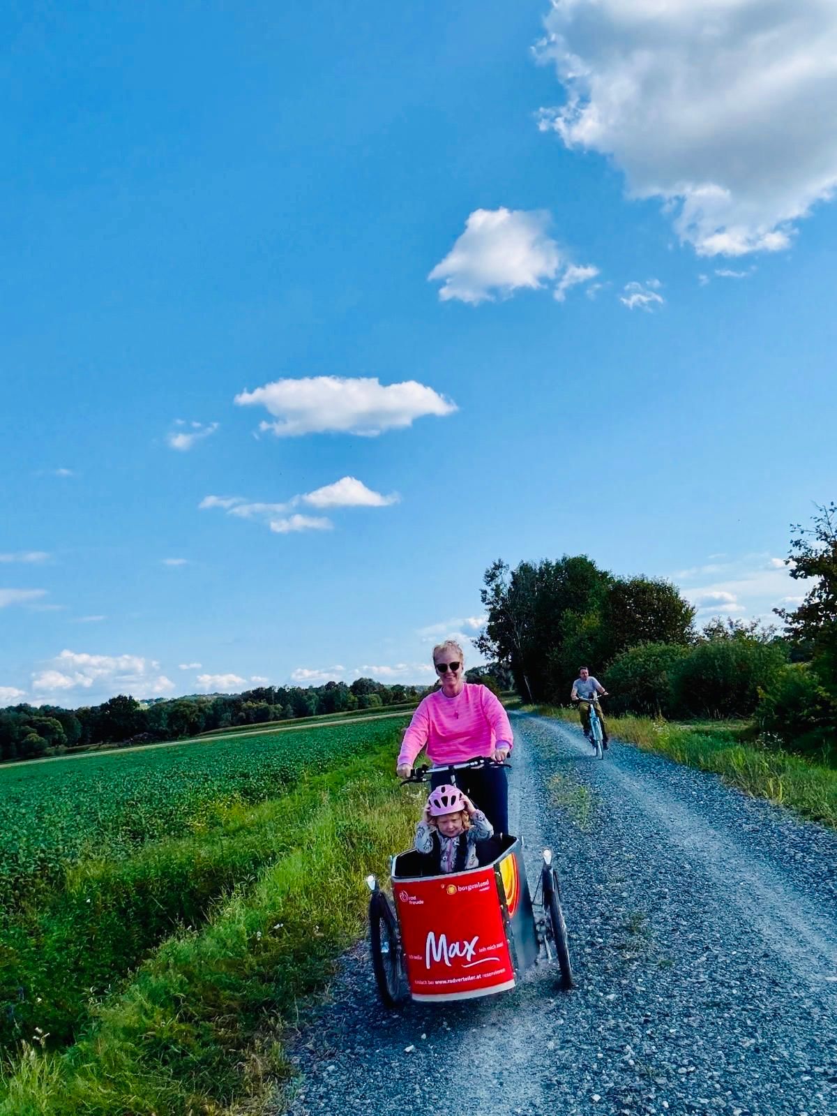Eine Frau fährt mit einem Kind in einem roten Max-Anhänger auf einer Kiesstraße unter einem blauen Himmel mit verstreuten Wolken.