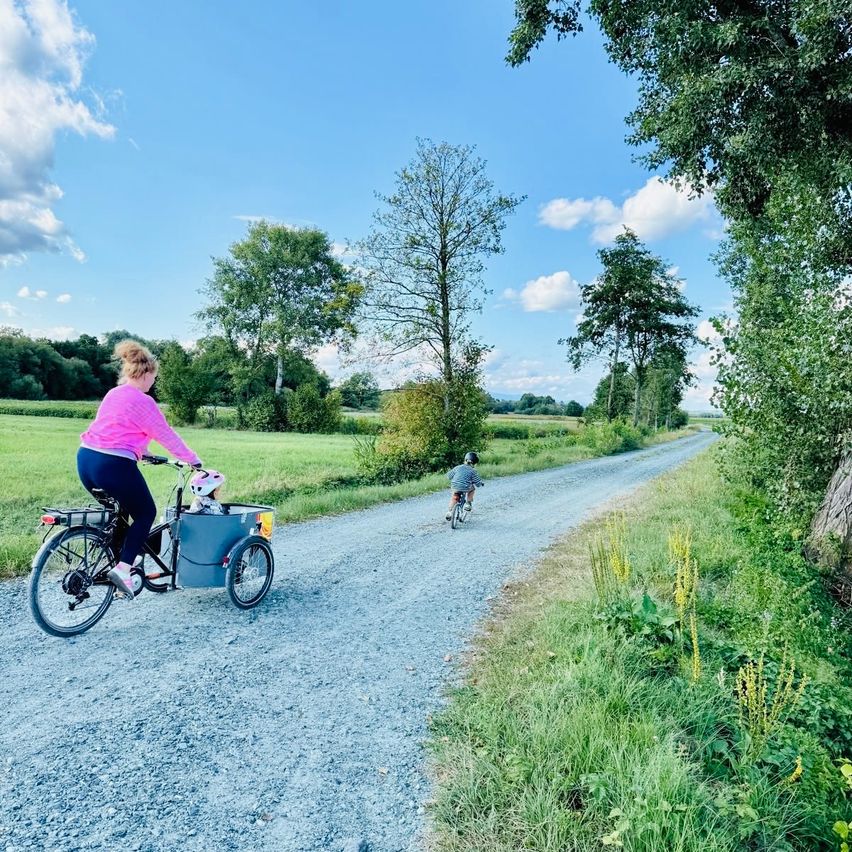 Eine Frau fährt mit einem Kind in einem Anhänger auf einem Schotterweg, der von grünen Feldern und Bäumen gesäumt ist. Ein Kind fährt hinter ihr auf einem Fahrrad.