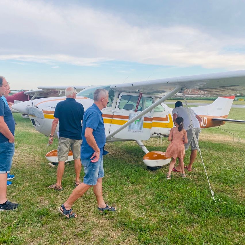 Eine Gruppe von Menschen steht um ein Flugzeug auf einem Grasfeld. Ein Mädchen klettert die Leiter des Flugzeugs hinauf.