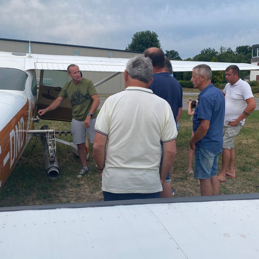 Eine Gruppe von Männern in Freizeitkleidung steht um ein kleines Flugzeug auf einem Grasfeld. Der Pilot steht in der offenen Tür des Flugzeugs.