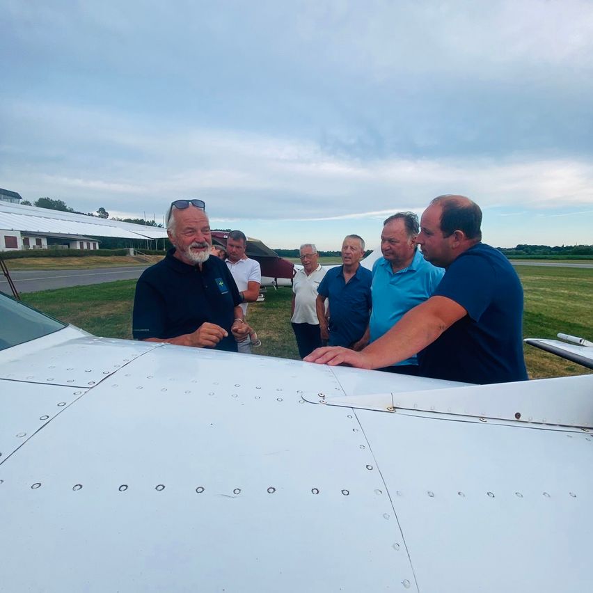 Sechs Männer stehen um ein weißes Flugzeug herum. Einer berührt den Flügel des Flugzeugs, während die anderen zusehen. Sie befinden sich auf einem Grasfeld bei bewölktem Himmel.