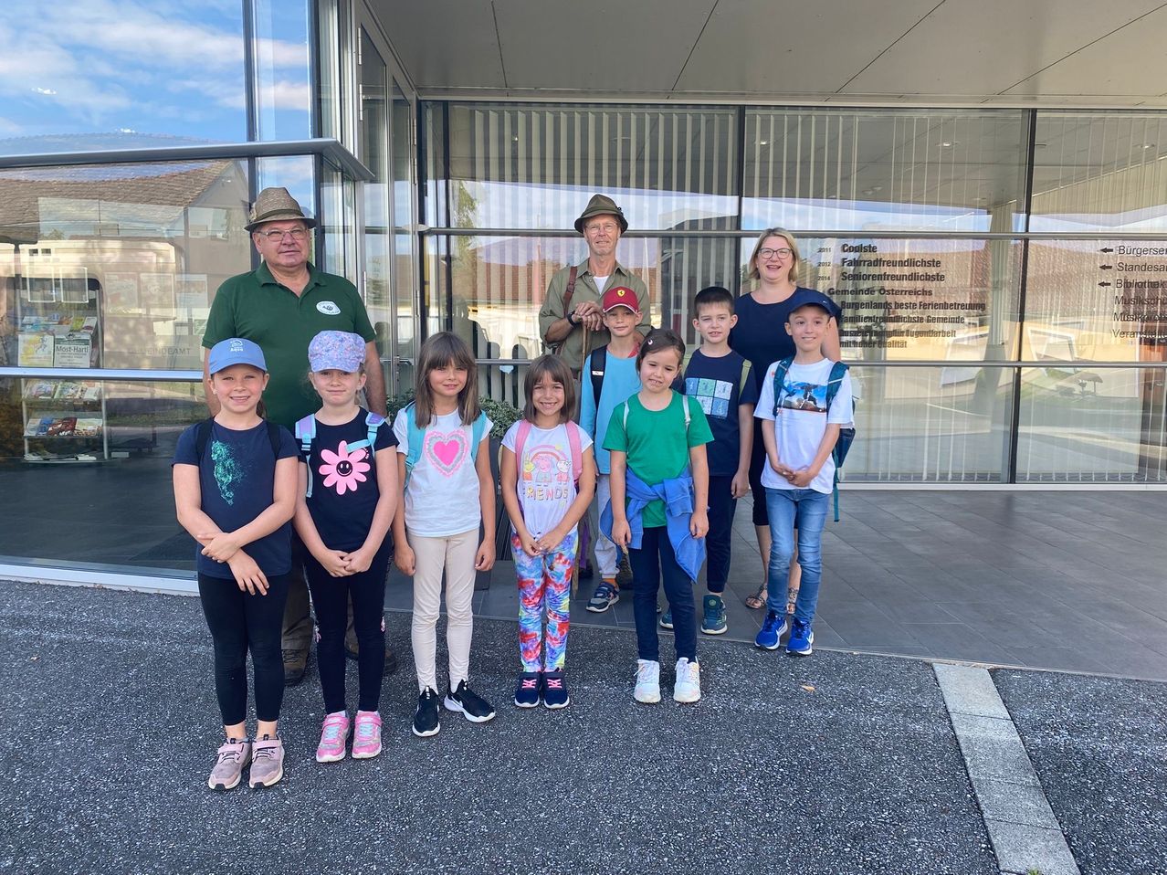 Eine Gruppe von Kindern und Erwachsenen steht vor einem Gebäude und lächelt für ein Foto. Die Erwachsenen tragen Hüte und Brillen, während die Kinder verschiedene Outfits tragen. Das Gebäude hat Glaswände und Türen, die die Umgebung reflektieren.