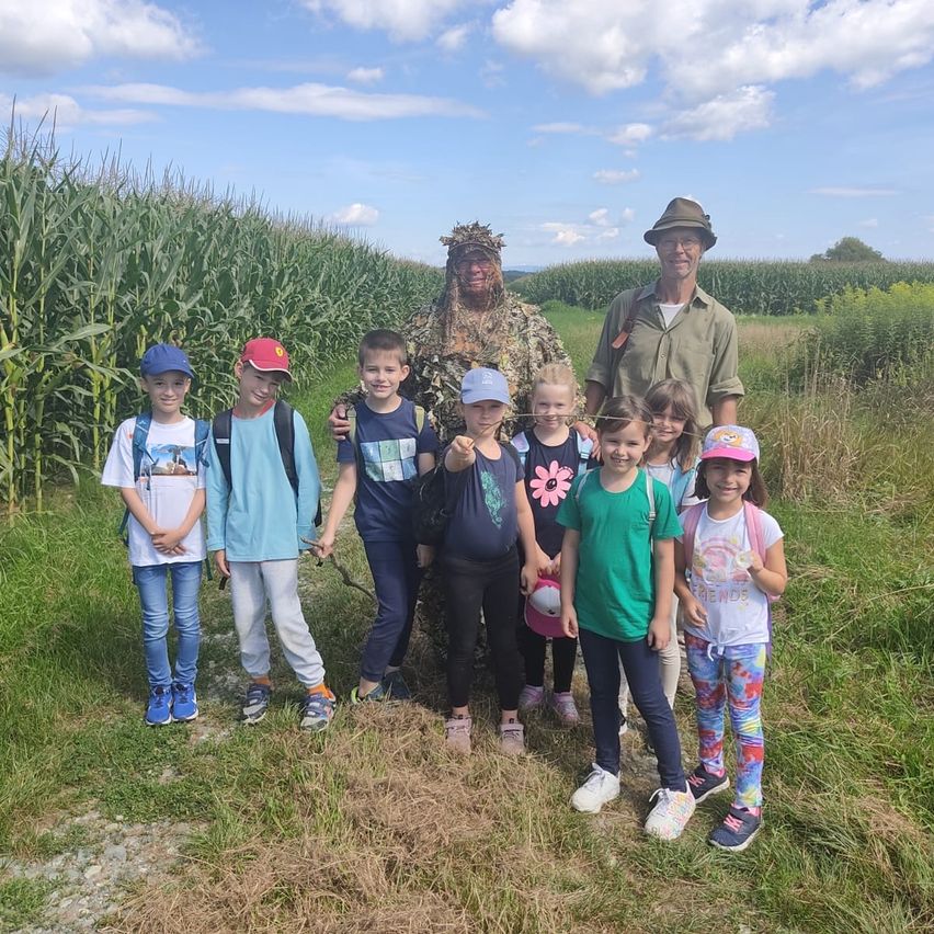 Eine Gruppe von Kindern mit zwei Erwachsenen in einem Maisfeld. Ein Erwachsener trägt Tarnkleidung, der andere ein grünes Hemd. Die Kinder stehen eng beieinander, einige lächeln.