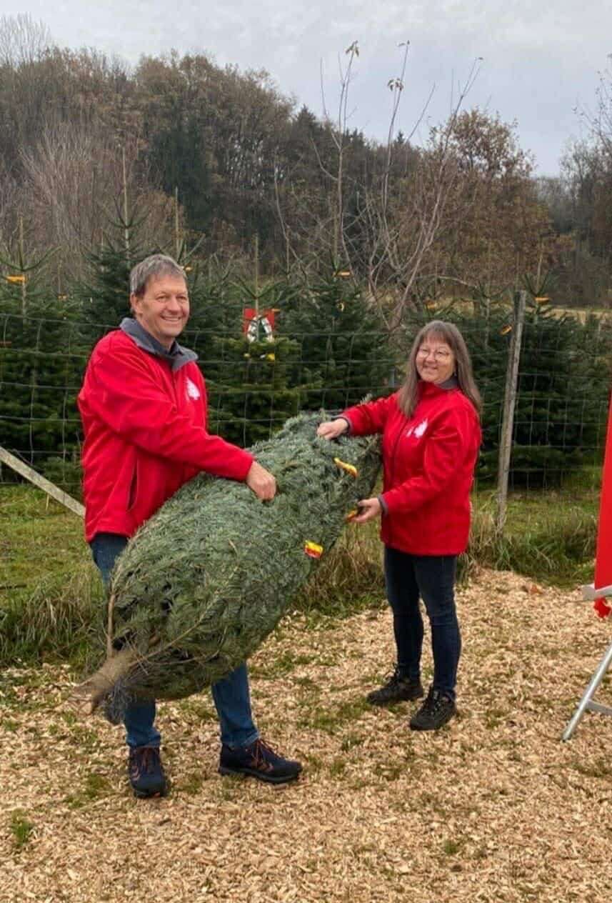 Ein Mann und eine Frau halten einen großen Weihnachtsbaum in einem Kiefernwald. Sie tragen beide rote Jacken und lächeln. Dahinter befindet sich ein Zaun mit Kiefernbäumen.