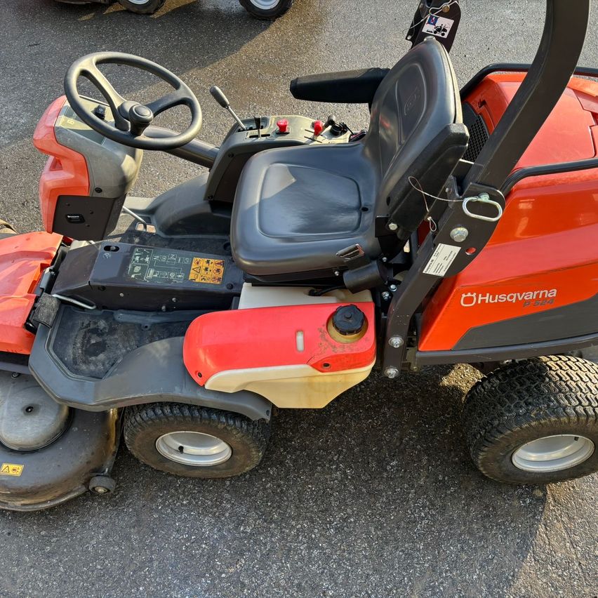 Ein roter und schwarzer Husqvarna Rasenmäher mit schwarzem Sitz steht auf einer gepflasterten Oberfläche. Er verfügt über ein Lenkrad, rote Knöpfe und einen Kraftstofftank.