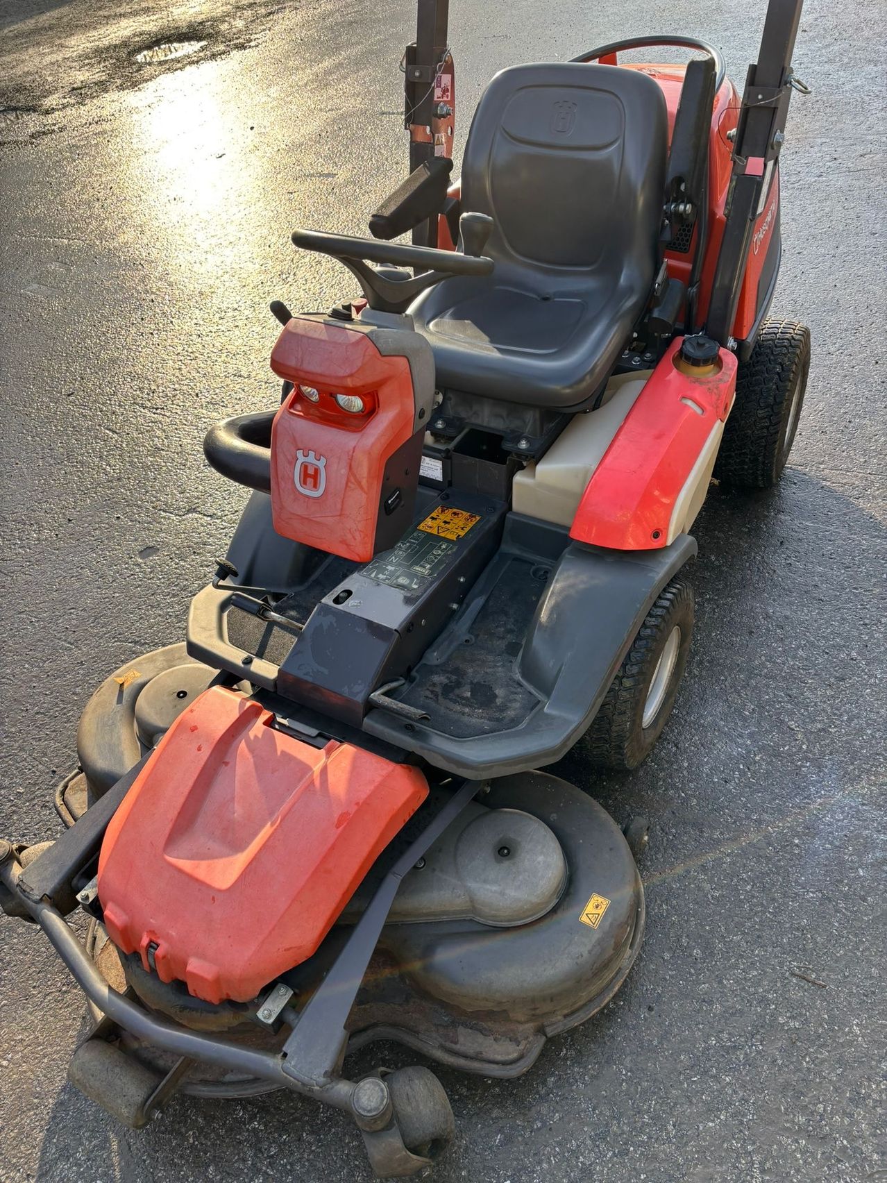 Ein roter und schwarzer Rasenmäher mit einer roten Abdeckung über dem Motor und zwei großen Rädern steht auf einer gepflasterten Oberfläche.