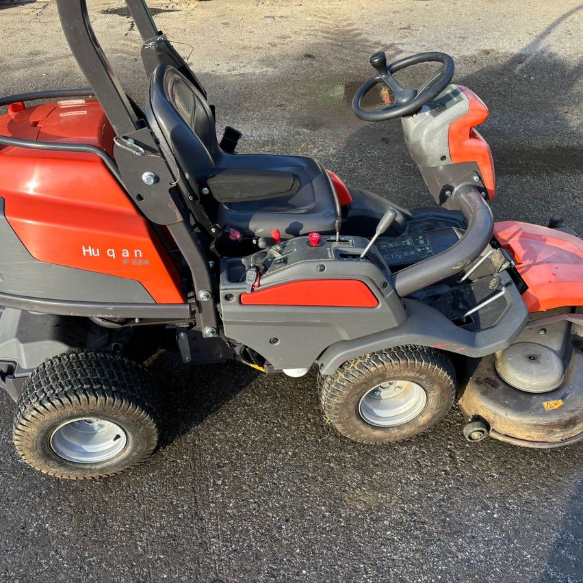 Ein orangefarbener und grauer Husqvarna-Rasenmäher mit zwei schwarzen Sitzen steht auf einer gepflasterten Oberfläche.