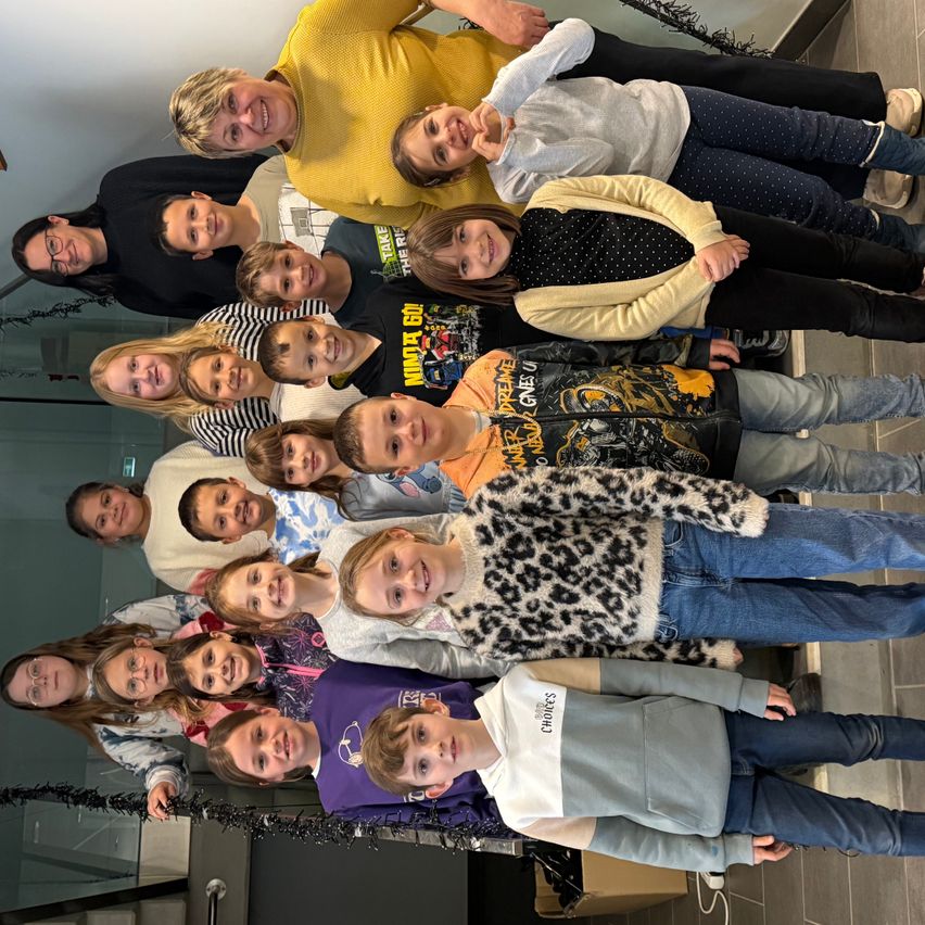 Eine Gruppe lächelnder Kinder und Erwachsener posiert für ein Foto. Einige tragen eine Brille, und sie haben verschiedene Kleidungsstücke. Eine Erwachsene trägt einen gelben Pullover, und ein Kind trägt einen Leoparden-Druck-Pullover. Sie stehen auf gefliesten Böden mit einer Treppe.