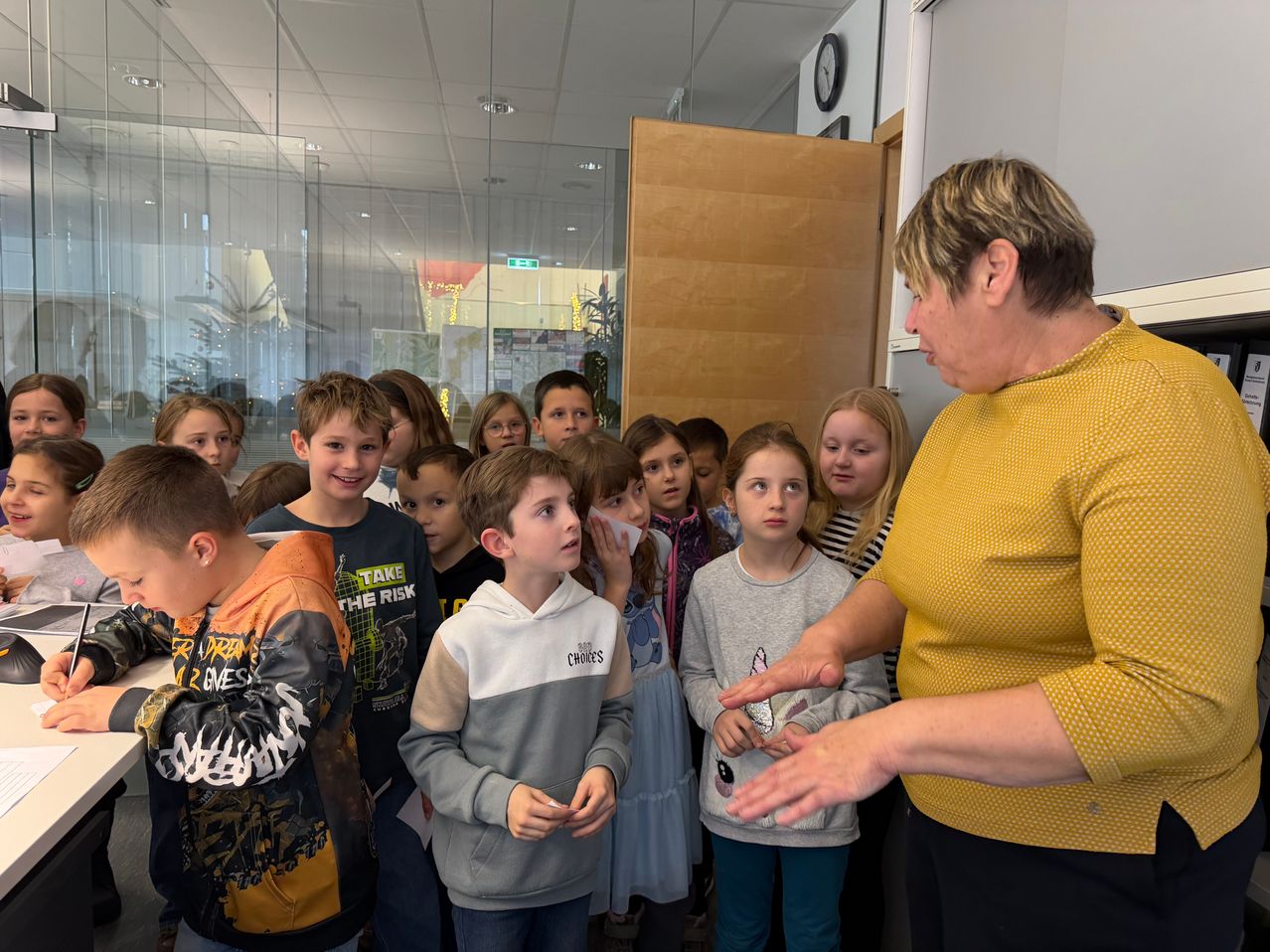 Eine Frau mit kurzen Haaren spricht mit einer Gruppe von Kindern in einem Flur. Die Kinder hören aufmerksam zu. Die Frau trägt ein gelbes Oberteil.