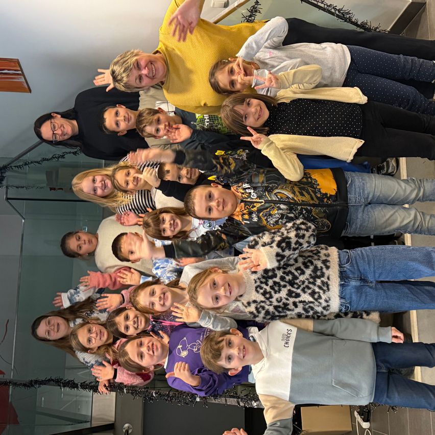 Eine Gruppe von Kindern und Erwachsenen posiert für ein Foto, lächelt und macht Gesten. Der Hintergrund ist eine Glaswand mit dekorativen Lichtern.