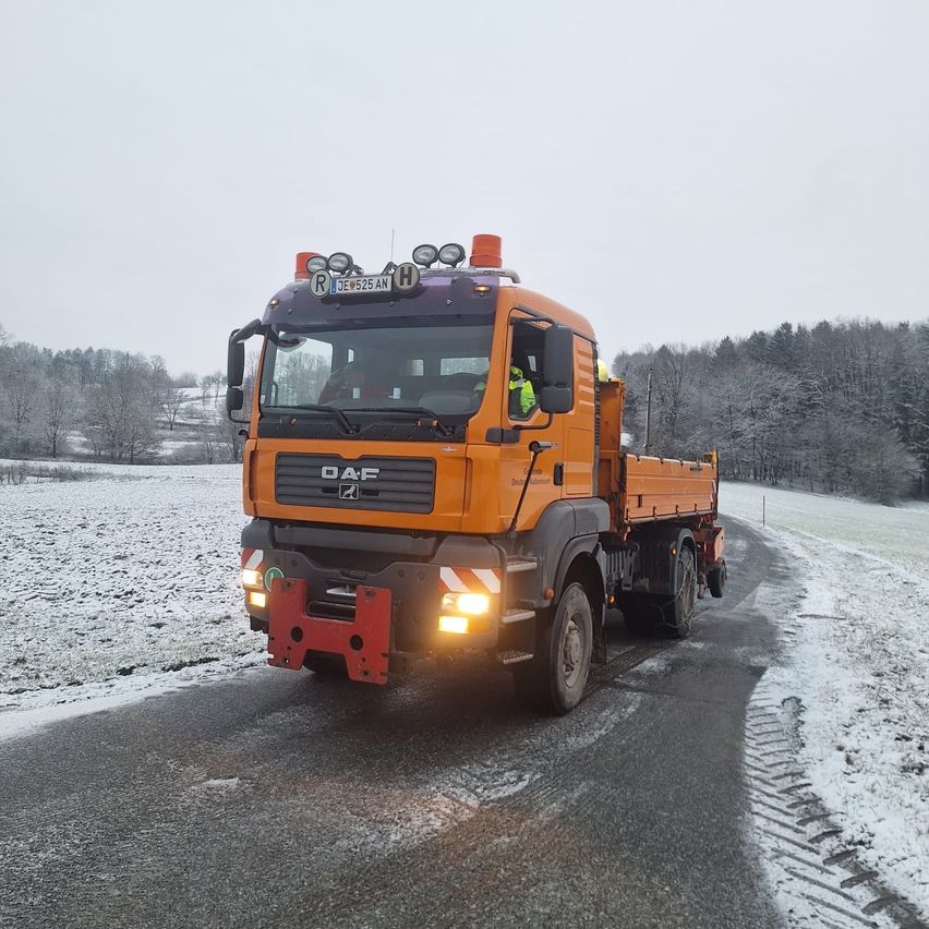 Ein orangefarbener Lastwagen mit einem Pflug fährt auf einer verschneiten Straße. Bäume sind schneebedeckt.